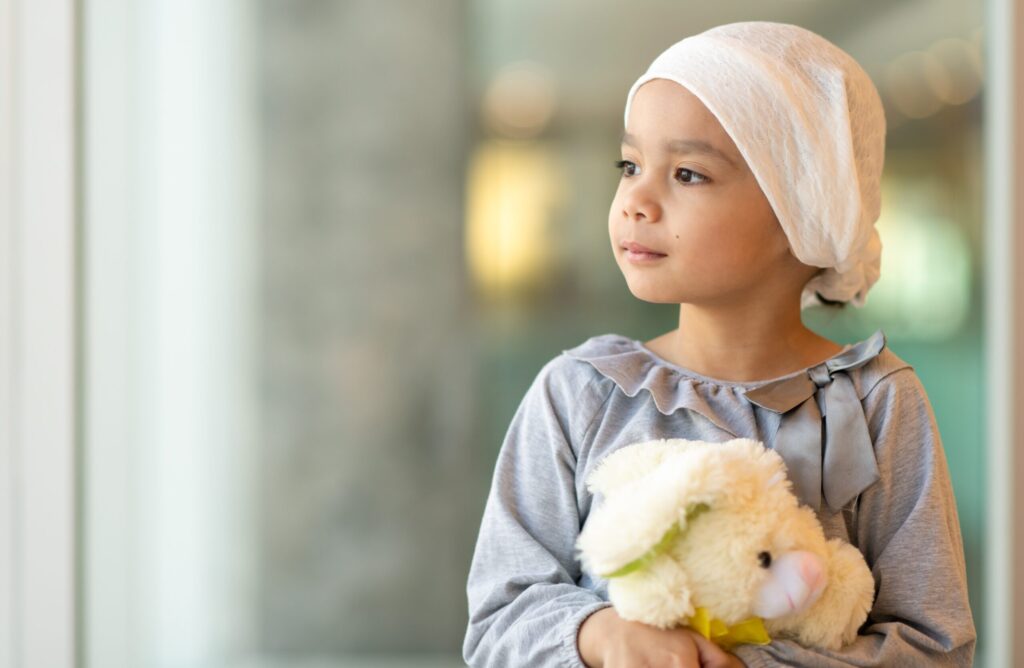 A beautiful little girl with cancer takes a break from treatment. She is standing near a large bay of windows in the hospital's corridor. The girl is wearing a headscarf and is holding a stuffed rabbit toy. She is looking out the window with a peaceful expression.